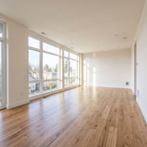 A bright, clean, completely empty living room of a modern apartment with large windows and beautiful hardwood floors, bathed in natural sunlight.